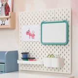 White wall-mounted pegboard with a teal-framed magnetic drawing board and containers holding colored cups and scissors.