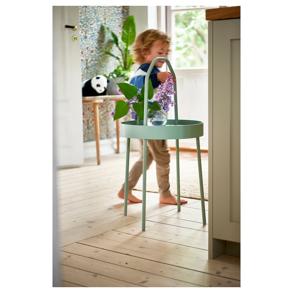 Child playing near a mint green round side table with a handle, holding purple flowers.