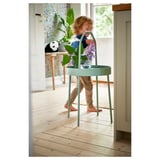 Child playing near a mint green round side table with a handle, holding purple flowers.