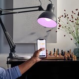 A person controls a lamp via smartphone app, changing its color, beside a chessboard and flowers.
