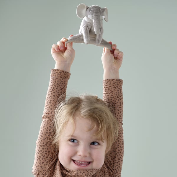 Child holds grey elephant plush toy smiling. From DJUNGELSKOG, recycled polyester, ages 12m+