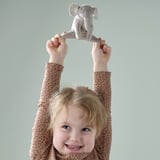 Child holds grey elephant plush toy smiling. From DJUNGELSKOG, recycled polyester, ages 12m+