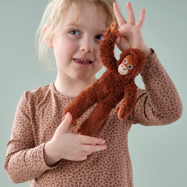 Blonde child smiling, holding a plush brown toy monkey with arms raised, both making peace signs.