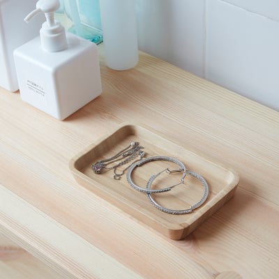 Bamboo jewelry tray with necklaces and bangles on wooden counter with soap and blue bottle.