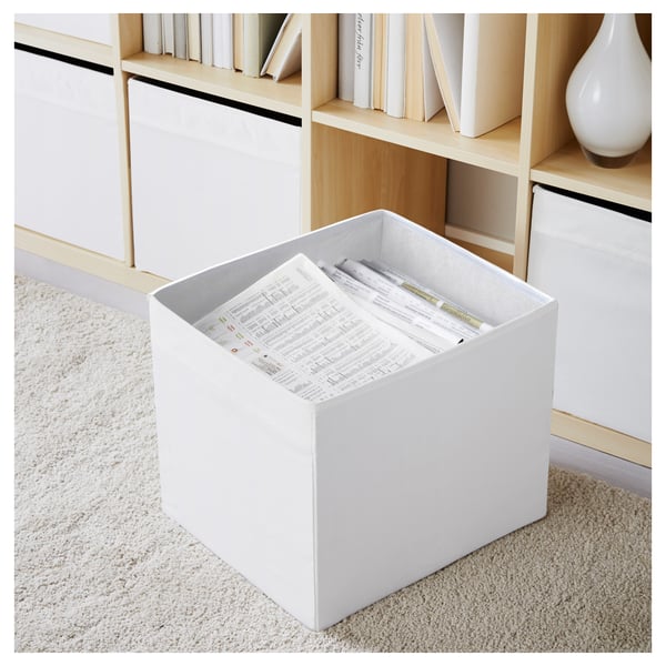 White storage box on carpet, filled with newspapers and pens, part of a wooden shelf unit.