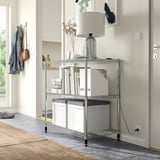 A modern entryway with a metallic 3-tier storage shelf holding white boxes, a lamp, and books. A grey jacket and bag hang nearby.