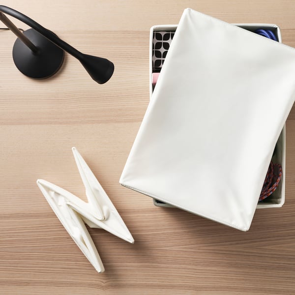 A white fabric lid for storage boxes on a wooden table, covering a container with objects inside.