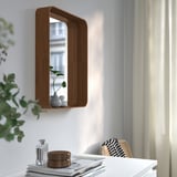 Wall-mounted mirror with shelf, reflecting window. Holds small vase with plants on shelf. Surrounded by white furniture and greenery.