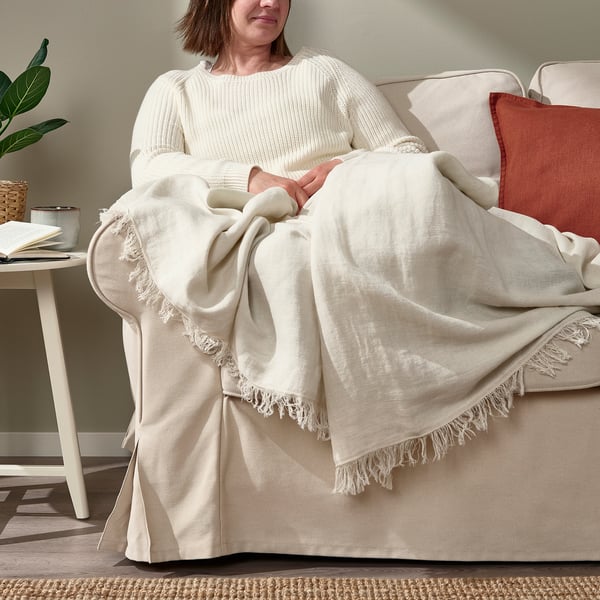 Woman sitting on beige couch with off-white DYTÅG throw blanket over legs, showing its soft texture and fringe ends.