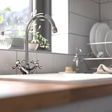 Chrome EDSVIK kitchen tap with high arc, plant, dish rack. Classic cross handles, white tiles, light countertop.