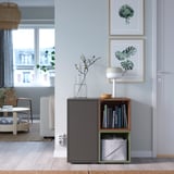 Modern living room with EKET grey storage unit displaying books and a lamp. Light wood floors and blue walls with large windows.