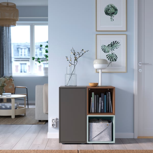 A gray EKET storage unit with open shelves and a door, holding books and a vase, next to a doorway.