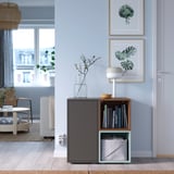A gray EKET storage unit with open shelves and a door, holding books and a vase, next to a doorway.