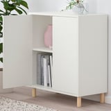 White EKET cabinet with two doors open, displaying books and a vase inside. It stands on wooden legs, with a plant on top.