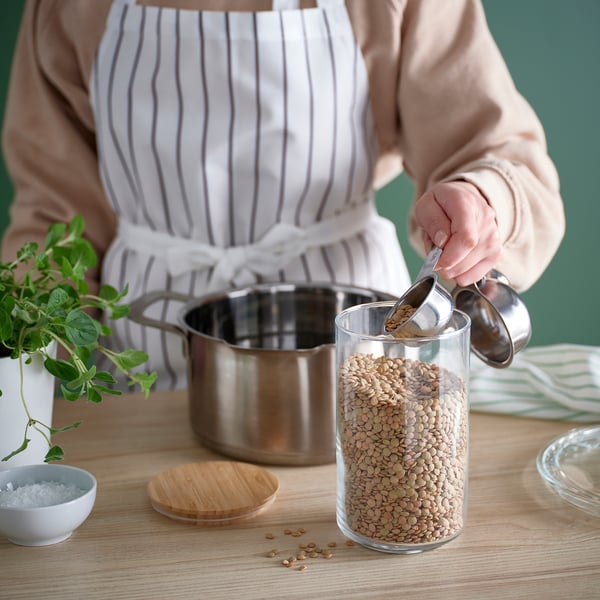 Woman measuring green lentils with a stainless-steel scoop from a transparent glass jar for cooking.