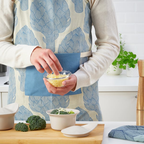 Person in apron grates cheese in kitchen with broccoli on counter.