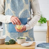 Person in apron grates cheese in kitchen with broccoli on counter.