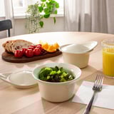 Table set with food: a bowl of salad, bread, fruits, and orange juice. A white ceramic dish, likely for baking and serving, is present on the table.