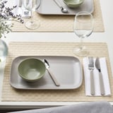 Table set with FÄRGKLAR plates, bowls, cutlery, and wine glasses on woven placemats.
