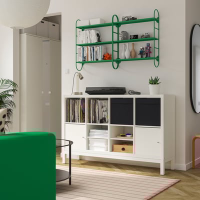 Green shelf with books, vases, cameras above cabinet with vinyl, books, printer. Green chair, striped rug below.