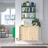 Modern wall shelf with books, decor, and plants; wooden cabinet below; nearby window and stool.