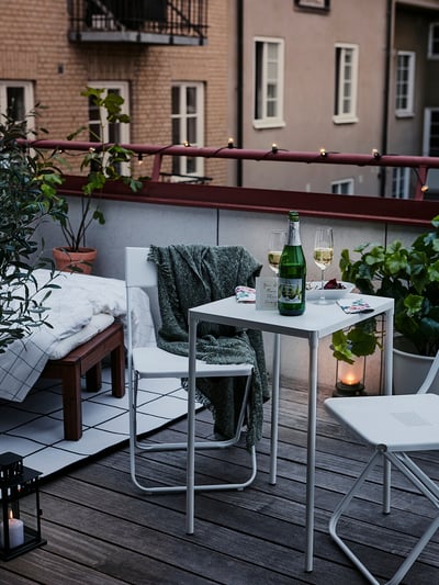 Cozy balcony with white FEJAN table, two chairs, blanket, and wine glasses. Wooden floor, potted plants, candles, and nearby building.