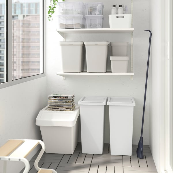 Organized storage area with white FILUR bins, some with lids, on shelves and floor; black push broom leans against wall.