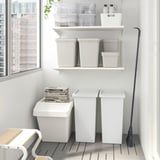 Organized storage area with white FILUR bins, some with lids, on shelves and floor; black push broom leans against wall.