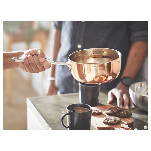 A rustic kitchen scene featuring FINMAT copper and stainless-steel saucepan, a black mug, and cookies on a wooden board.