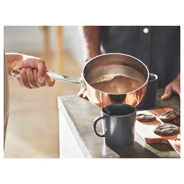 Hand holds shiny FINMAT saucepan over mug, chocolate cookies nearby on wet counter.