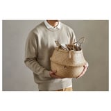 Person holding woven seagrass basket. Neutral beige tones, handles can be turned up or down.