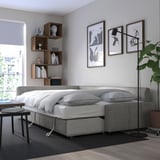 Modern bedroom with gray FRIHETEN sofa-bed, white bedding, and wooden bookshelf. Natural light from window, adjacent plant, and wall art.