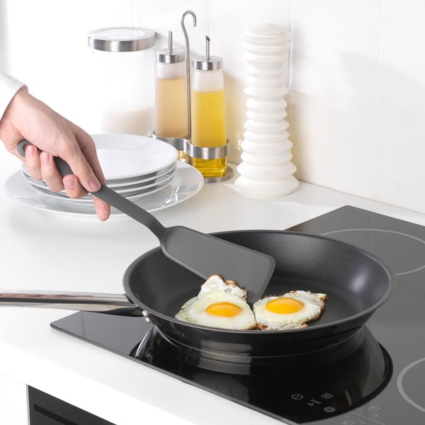 A hand uses a grey spatula to flip a fried egg in a black pan on a white stove.