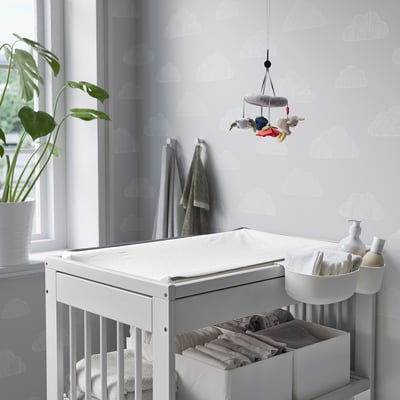 Baby changing table GULLIVER, white, wood, rectangular, with hanging mobile. Positioned near window, smaller than plant.