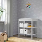 Baby changing table GULLIVER in white, fits in corner. Has shelves with storage boxes. Soft toys in basket beside. Pleasant nursery setup.