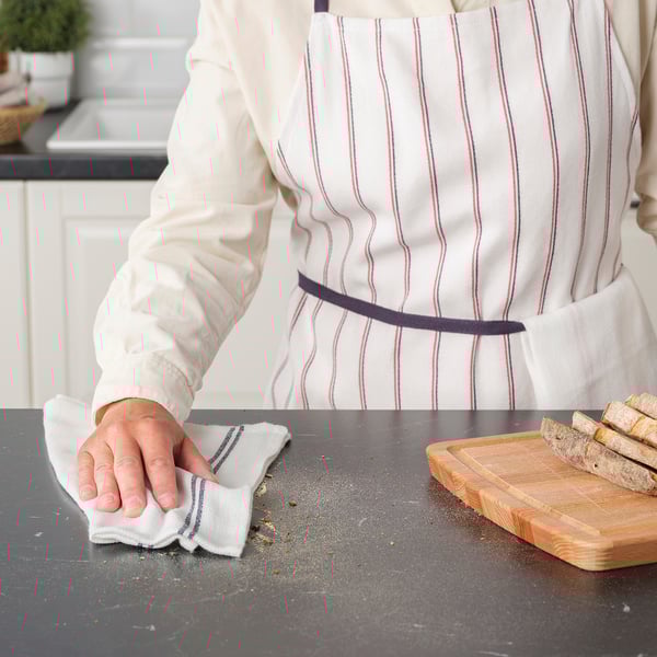 Person wearing apron cleans kitchen counter with tea towel, beside cutting board with bread slices.