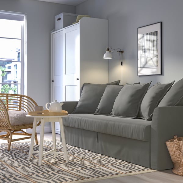 Modern living room with HOLMSUND sofa-bed, side table, woven chair, and rug. Sofa against wall with cushions and light above. Teapot on round table.