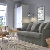 Modern living room with HOLMSUND sofa-bed, side table, woven chair, and rug. Sofa against wall with cushions and light above. Teapot on round table.