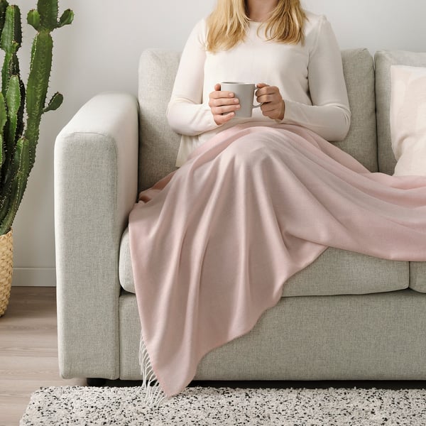 Woman sitting on sofa holding mug, wrapped in a soft pink HOLMVI blanket with fringes, providing warmth and comfort.