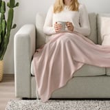 Woman sitting on sofa holding mug, wrapped in a soft pink HOLMVI blanket with fringes, providing warmth and comfort.