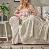 Woman sitting on beige chair, blanket over legs, holding coffee cup.