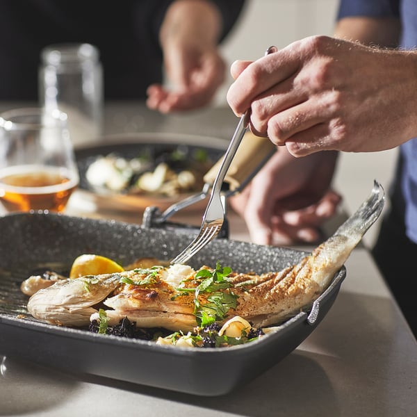Person eating grilled fish in a non-stick pan with comfortable handles, set on a table with drinks and another dish.