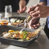 Person eating grilled fish in a non-stick pan with comfortable handles, set on a table with drinks and another dish.