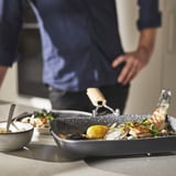 A person is cooking seafood in a non-stick, gray skillet with a lemon and herbs. The pan sits on a countertop, alongside a bowl with suspension sauce.