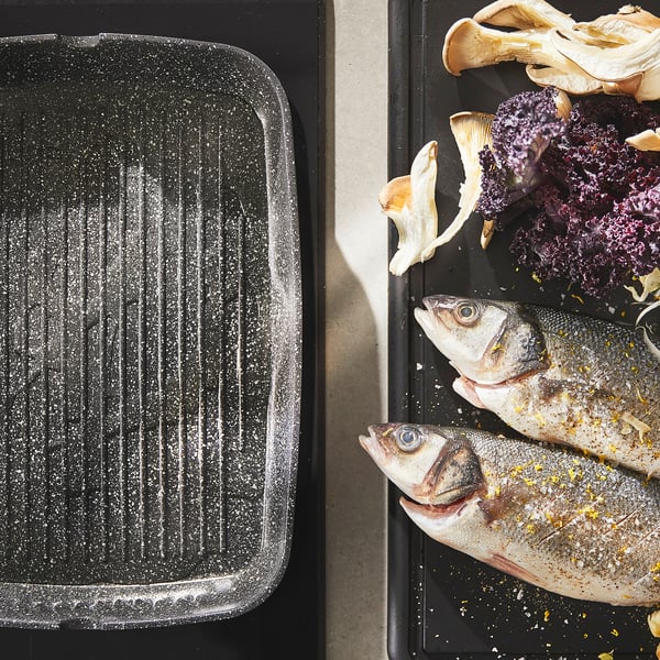 Two black cookware trays, one empty, the other with seasoned fish, vegetables ready for cooking.