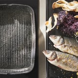 Two black cookware trays, one empty, the other with seasoned fish, vegetables ready for cooking.