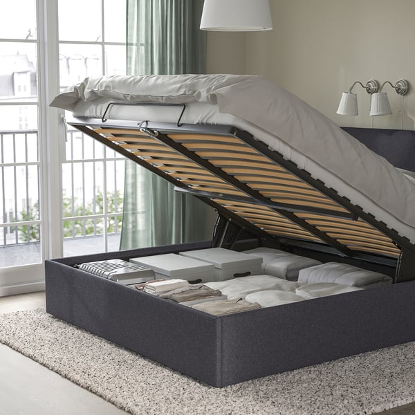 Bed with lifted storage compartment, showing organized blankets and pillows inside. Grey upholstered frame with slatted headboard.