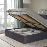 Bed with lifted storage compartment, showing organized blankets and pillows inside. Grey upholstered frame with slatted headboard.