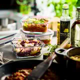 Glass food containers with lids holding colorful salads on a rustic table with a pan and herbs.