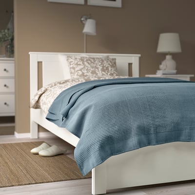 Blue cotton bedspread on large white bed, with pillows, lamp, and slippers nearby.
